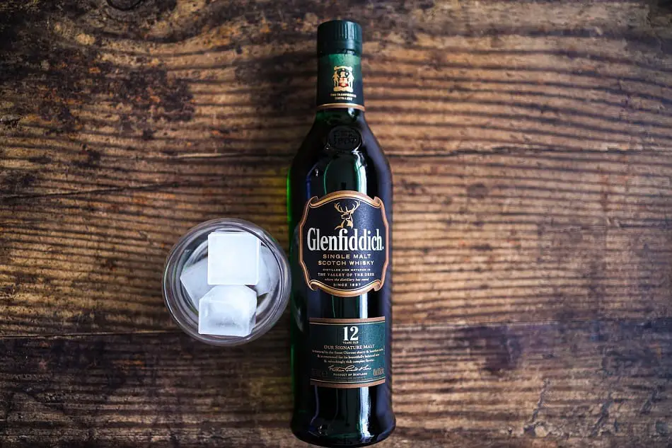 A glass with ice cubes and a bottle of 12 Year Old Glenfiddich lying on a wooden table