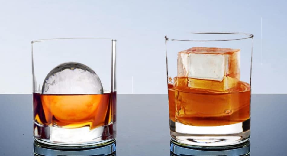 A glass of whiskey with large ice sphere and a glass of whiskey with a large ice cube