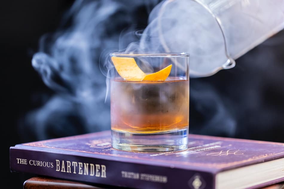 A smoking whiskey cocktail resting on the book 'The Curious Bartender'